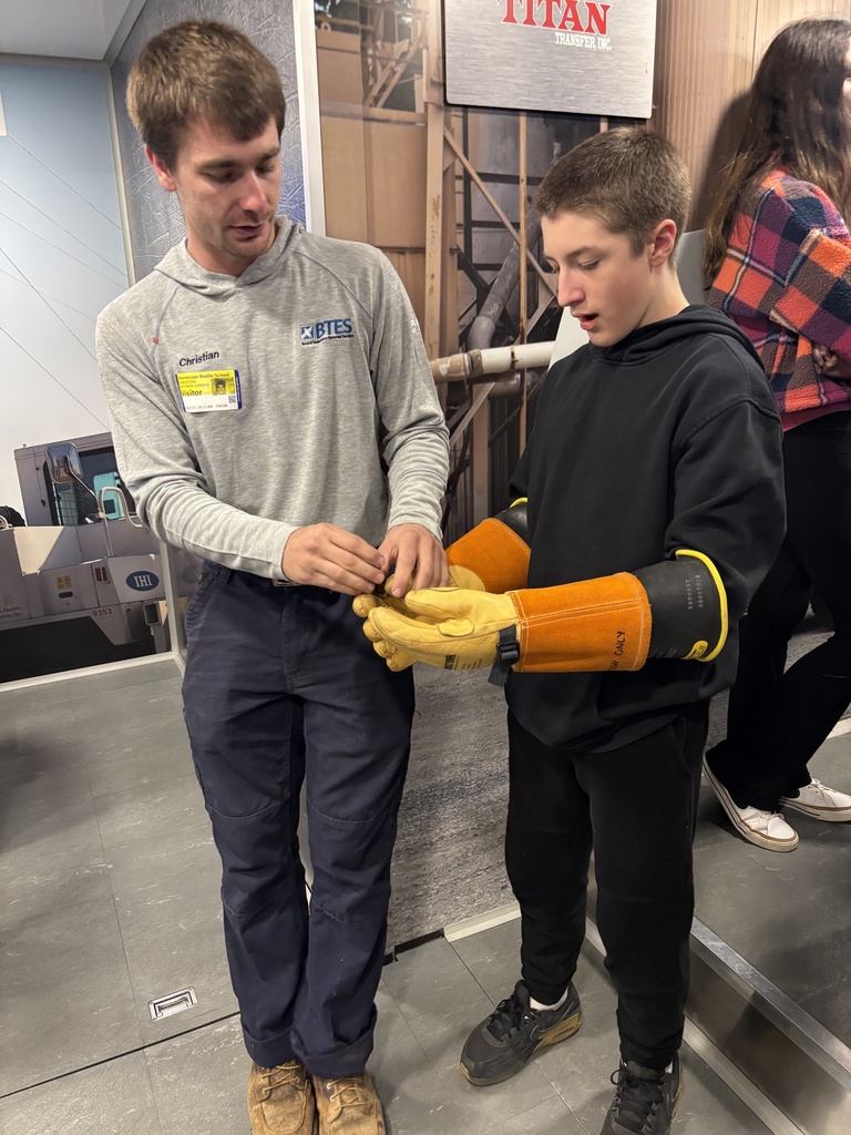 A BTES apprentice lineman helps a student try on protective lineman gloves inside the Be Pro Be Proud mobile workshop. The student looks at the gloves while the apprentice explains how they are used.