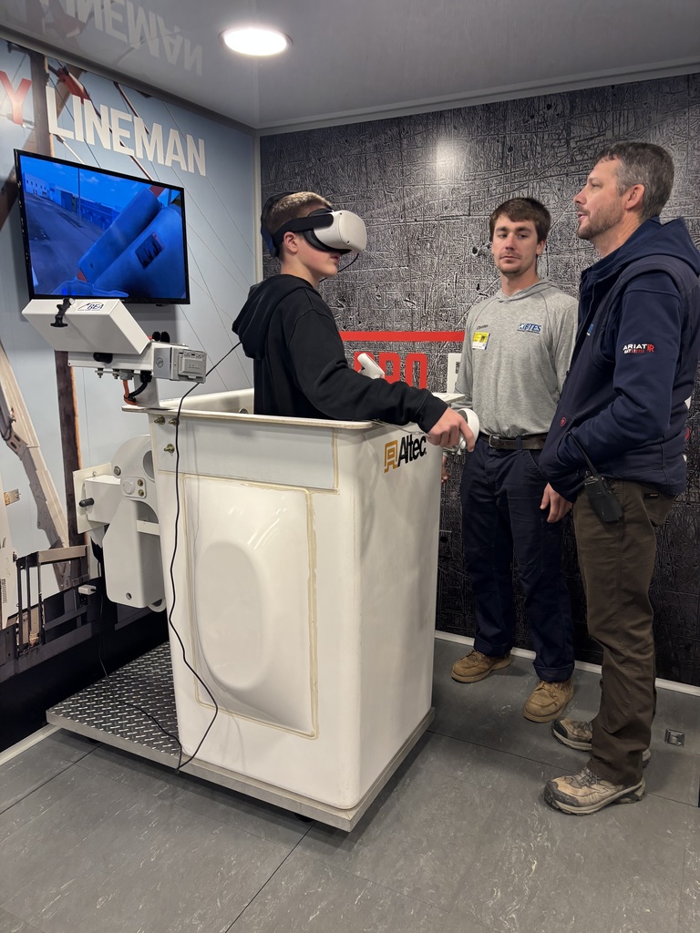 A student stands in a lineman bucket simulator wearing a VR headset and operating handheld controls while two BTES linemen observe and guide him.