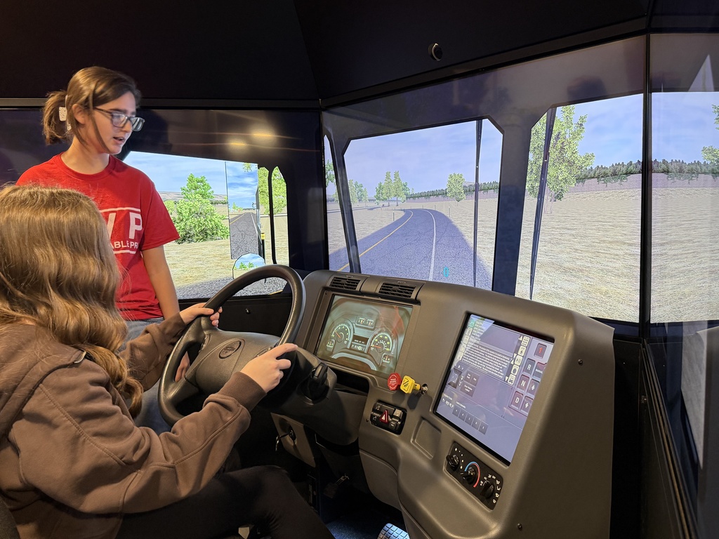 A Tennessee High School CTE student in a red shirt coaches a younger student who is seated at a commercial driving simulator, steering through a virtual roadway displayed on large curved screens.