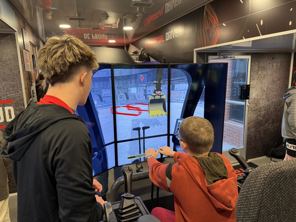 Two students work together on a heavy-equipment simulator, using joystick controls to operate a virtual excavator displayed on three large screens.