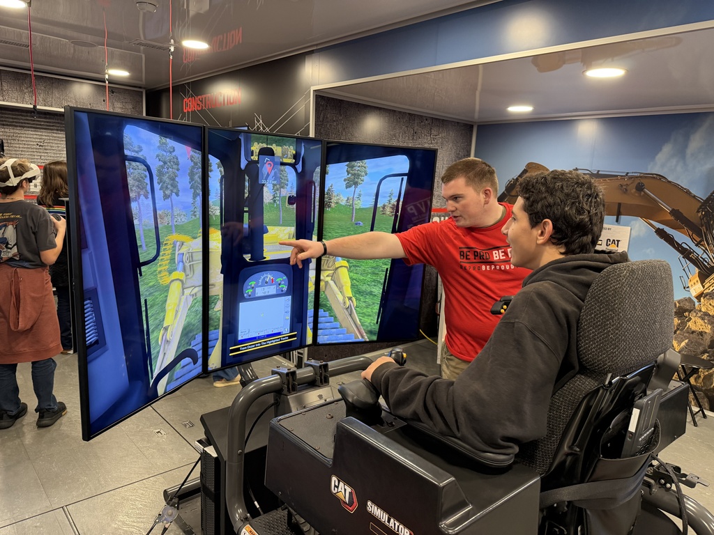 A Be Pro Be Proud staff member demonstrates a heavy-equipment simulator, pointing to the multi-screen display as a student in the operator seat uses the controls.