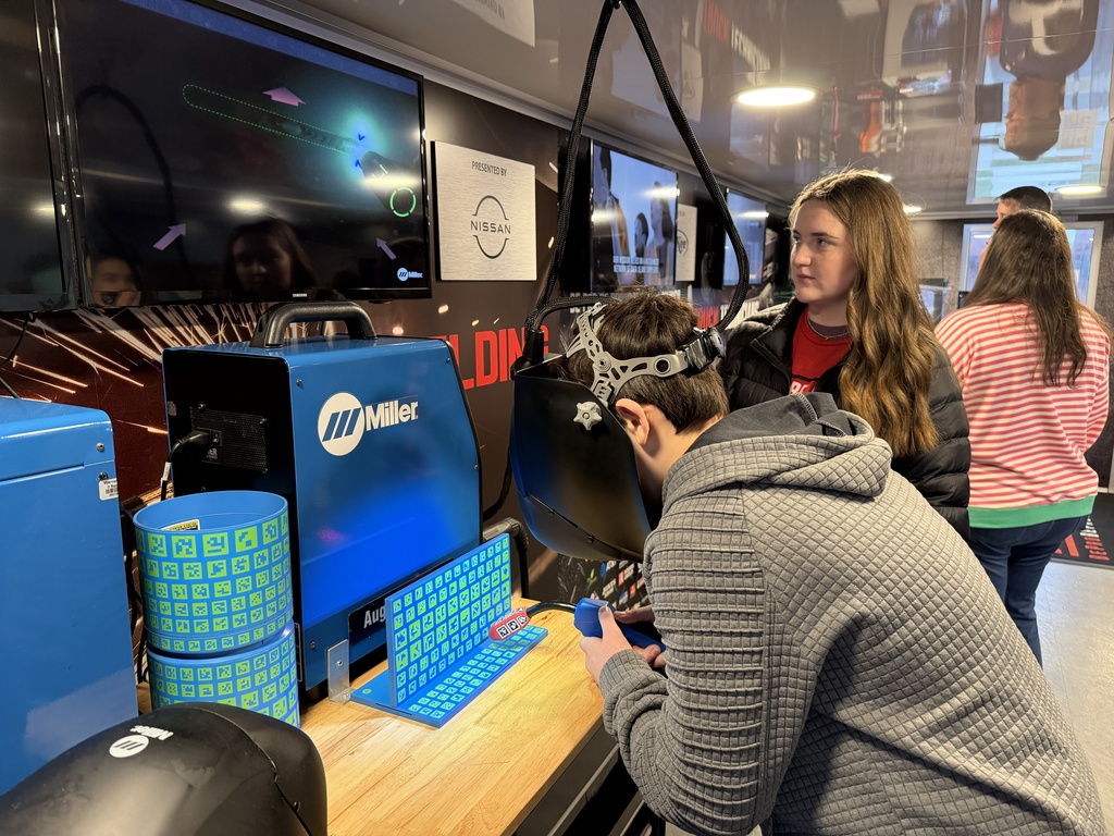 A student wearing a welding helmet practices on the virtual welding simulator while a Tennessee High CTE volunteer stands beside him. Welding equipment and an instructional screen are visible in the background.