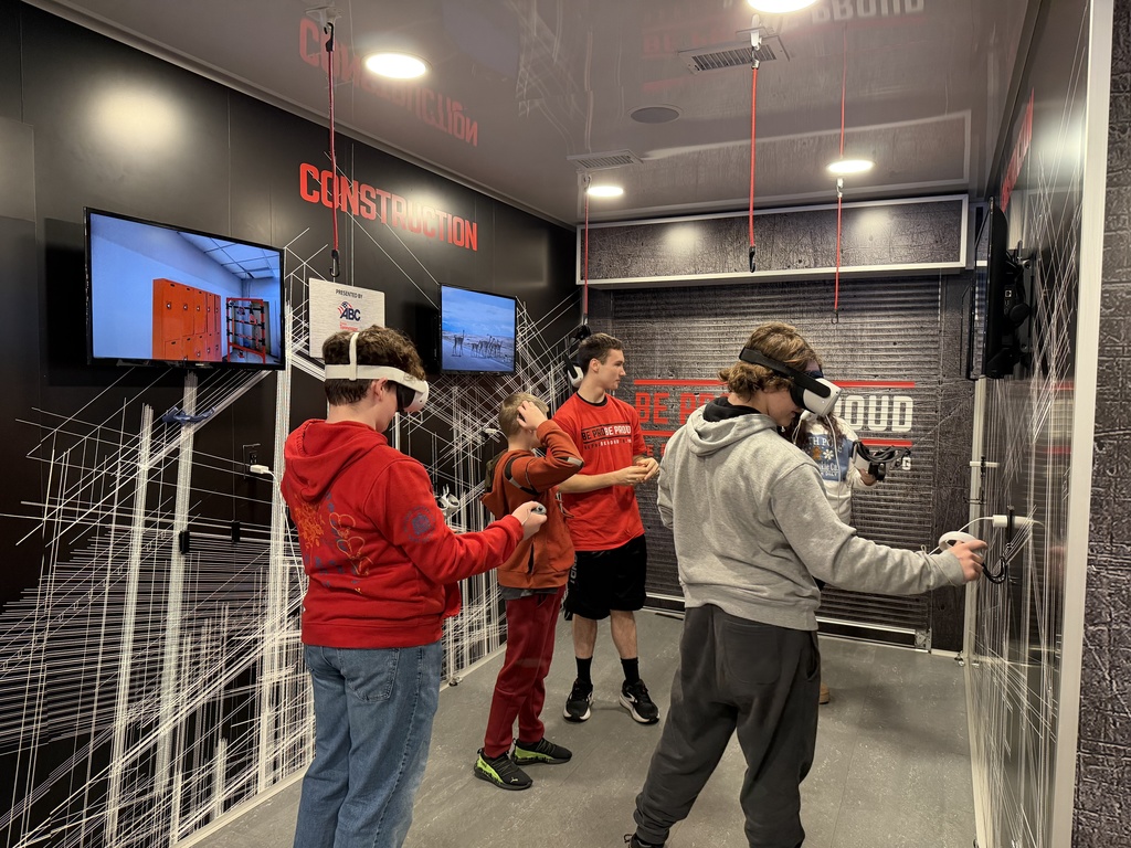 A group of middle school students inside the Be Pro Be Proud mobile workshop use virtual-reality construction simulators. Two students wear VR headsets while others watch and wait their turn, surrounded by construction-themed graphics and screens.