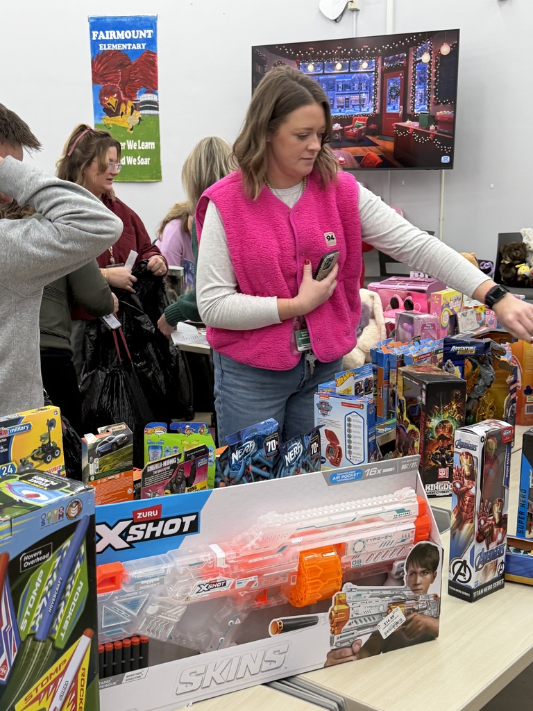 School counselors sort through donated toys in a room filled with tables of items. A woman in a bright pink vest stands in the foreground selecting a toy. Colorful boxed toys, including large Nerf and action-figure sets, fill the table in front of her.