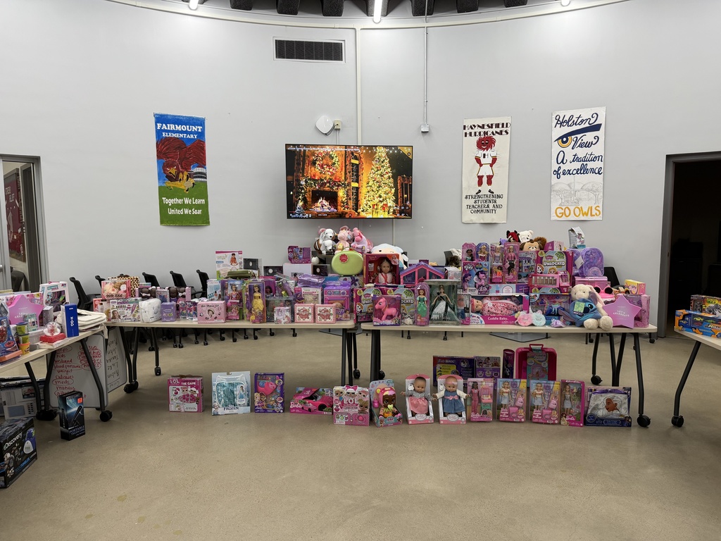 Rows of tables are filled with donated toys—dolls, playsets, stuffed animals, and children’s gift items—arranged in a school room decorated with banners for local elementary schools. A large TV on the back wall displays a Christmas fireplace scene.