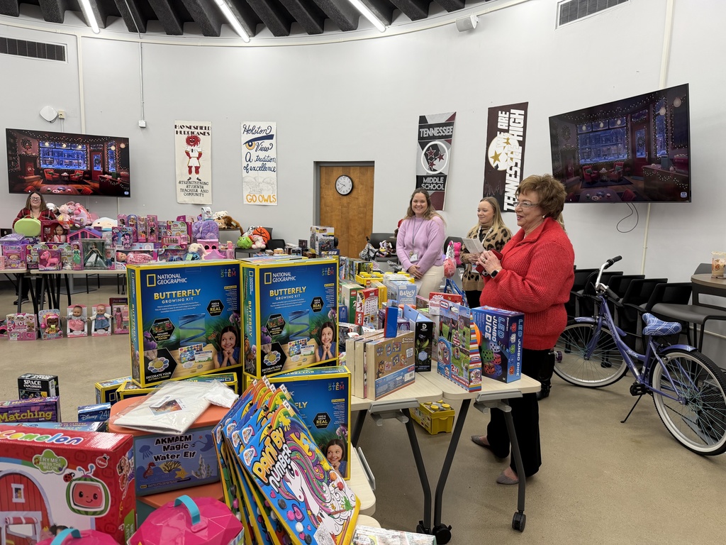 Several adults stand behind tables full of toys as one woman in a red sweater addresses the group. The room is lined with school banners, bicycles, and displays of STEM kits and children’s games. A TV screen behind them shows a decorated Christmas room.