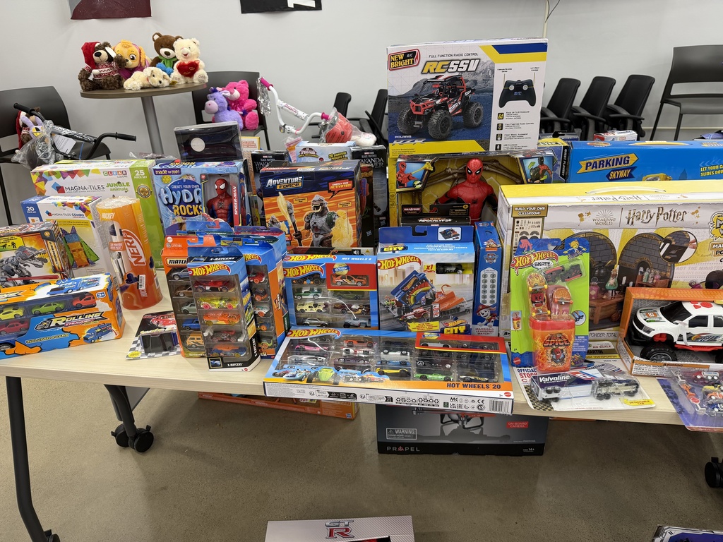 A close-up view of a table packed with toys, including Hot Wheels sets, Nerf products, remote-control cars, action figures, and various boxed playsets. Stuffed animals and bicycles are visible in the background.