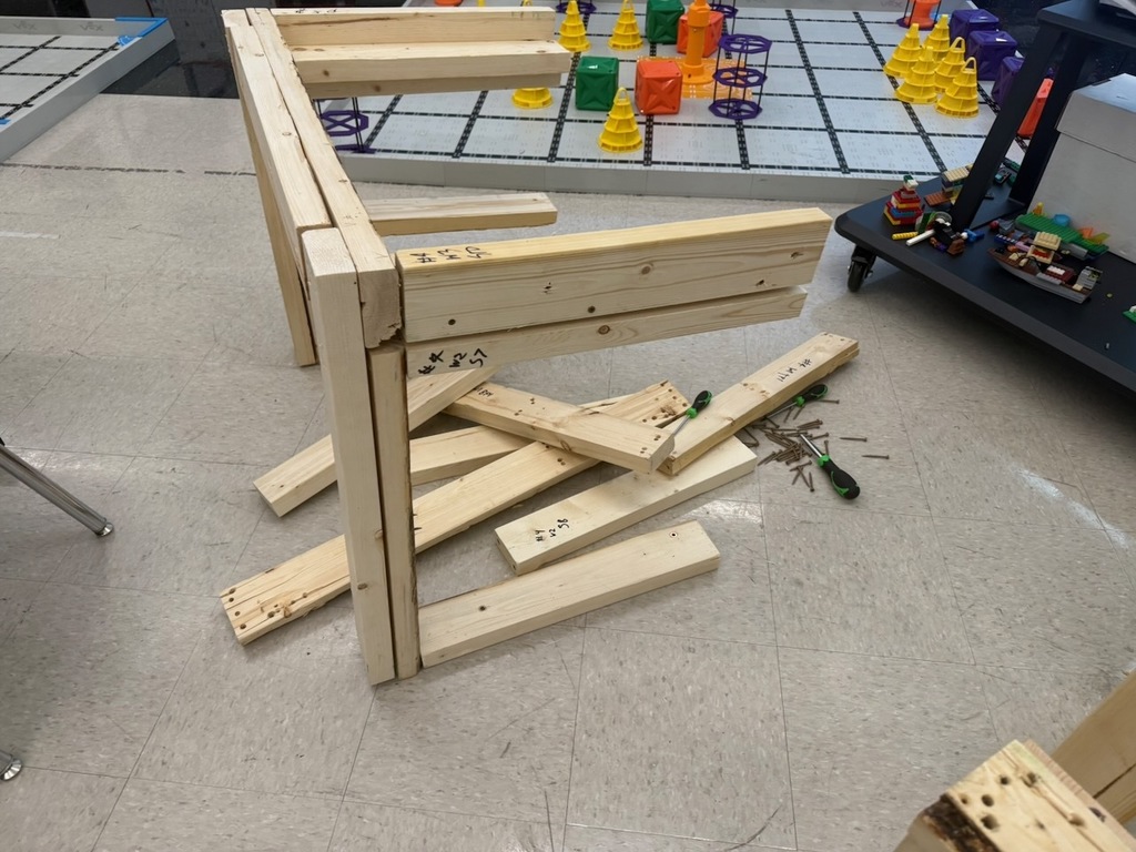 Partially assembled wooden frames and loose boards are scattered on the classroom floor near robotics equipment.