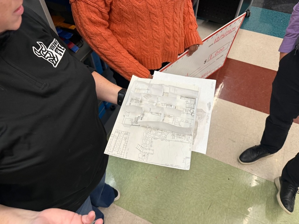 A teacher holds a student-made paper model and floorplan sketch, showing a detailed layout of rooms and structures. Several adults stand nearby on a multicolored tile floor, including one holding an oversized check in the background.