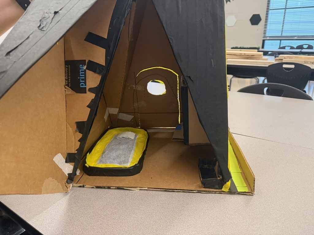 A small student-built cardboard structure resembling a triangular shelter is displayed on a classroom table. Inside, there is a handmade bed with yellow and black trim, a cutout window, and taped details along the walls.