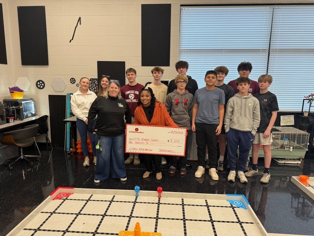 A group of Tennessee Middle School students stand with teacher Catherine Lamie and a Strongwell representative, who is holding an oversized check for $1,000. A robotics field is visible in front of them.