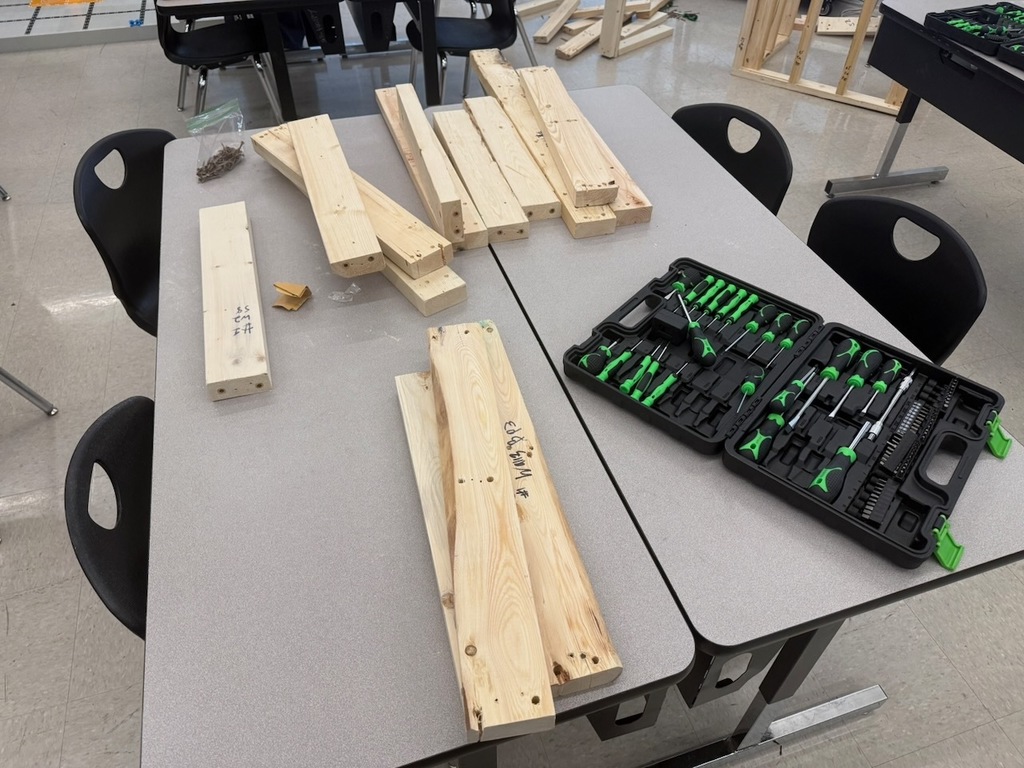 A classroom table covered with neatly arranged wooden boards, screws, and open toolkits with green-handled tools.