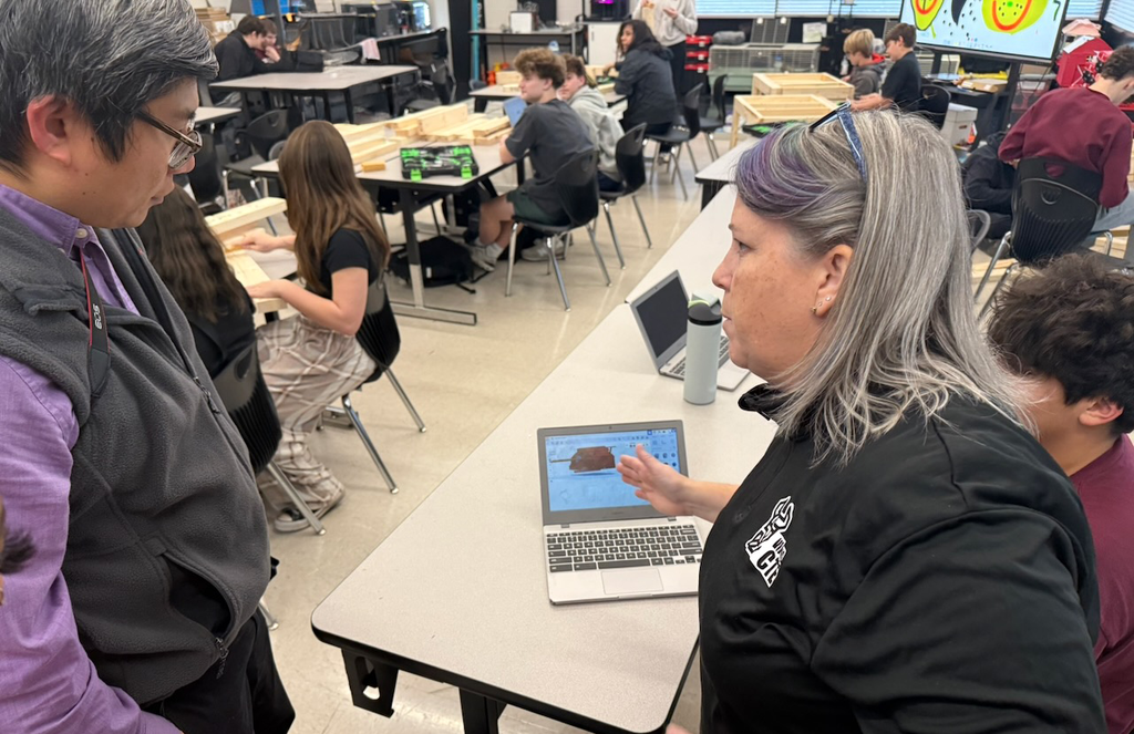 Teacher Catherine Lamie speaks with a visitor while showing a 3D model on a laptop; behind them, students work with wood pieces and tools at tables around the classroom.