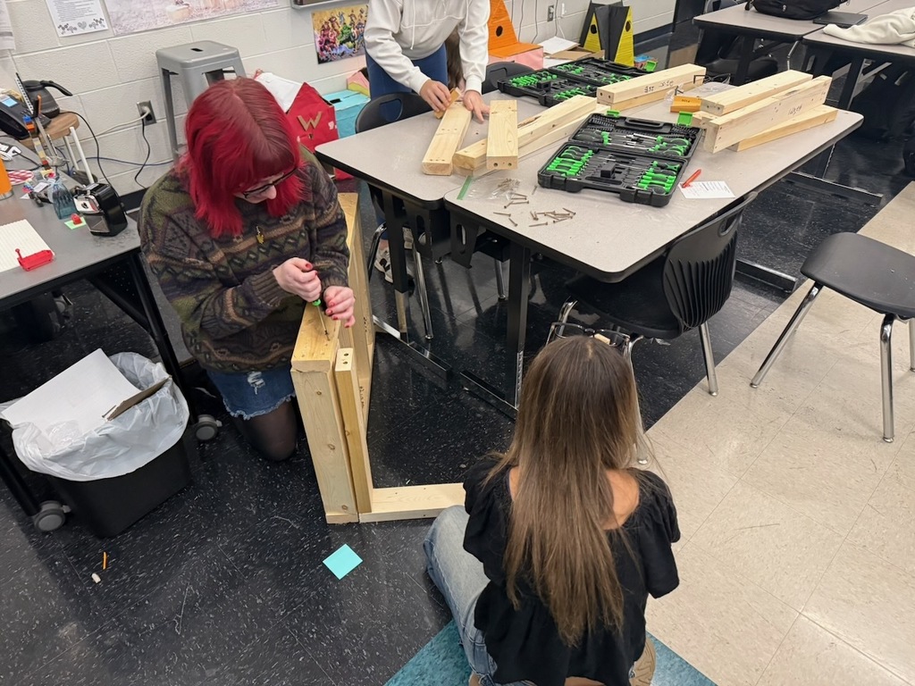 Students kneel on the floor assembling a wooden frame using screws and handheld tools. Pieces of lumber, toolkits, and classroom tables surround them.