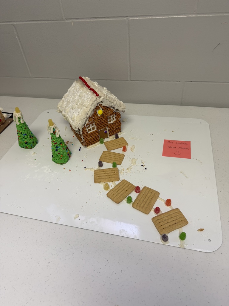 A collection of student-created gingerbread displays set on tables, featuring creatively decorated houses and scenes made from graham crackers, icing, candy, and small props. Designs include colorful candy roofs, snowy icing paths, miniature trees, a drive-in movie scene, a dinosaur-themed display, and humorous handwritten labels. The exhibits showcase imagination, craftsmanship, and festive holiday spirit.