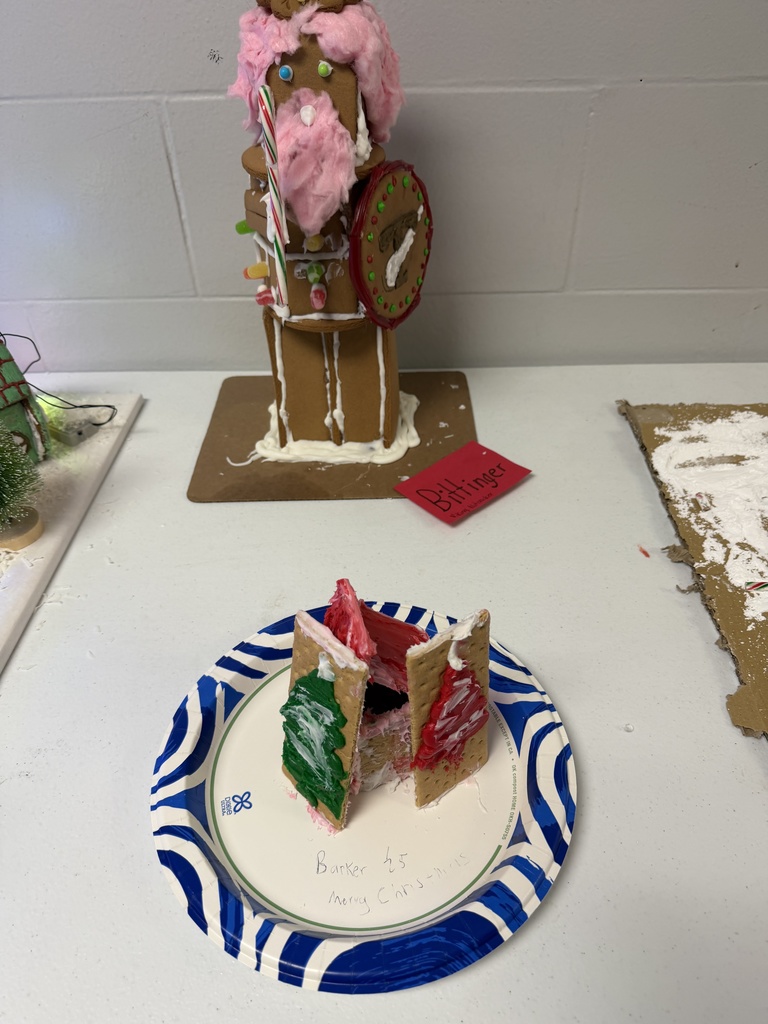 A collection of student-created gingerbread displays set on tables, featuring creatively decorated houses and scenes made from graham crackers, icing, candy, and small props. Designs include colorful candy roofs, snowy icing paths, miniature trees, a drive-in movie scene, a dinosaur-themed display, and humorous handwritten labels. The exhibits showcase imagination, craftsmanship, and festive holiday spirit.