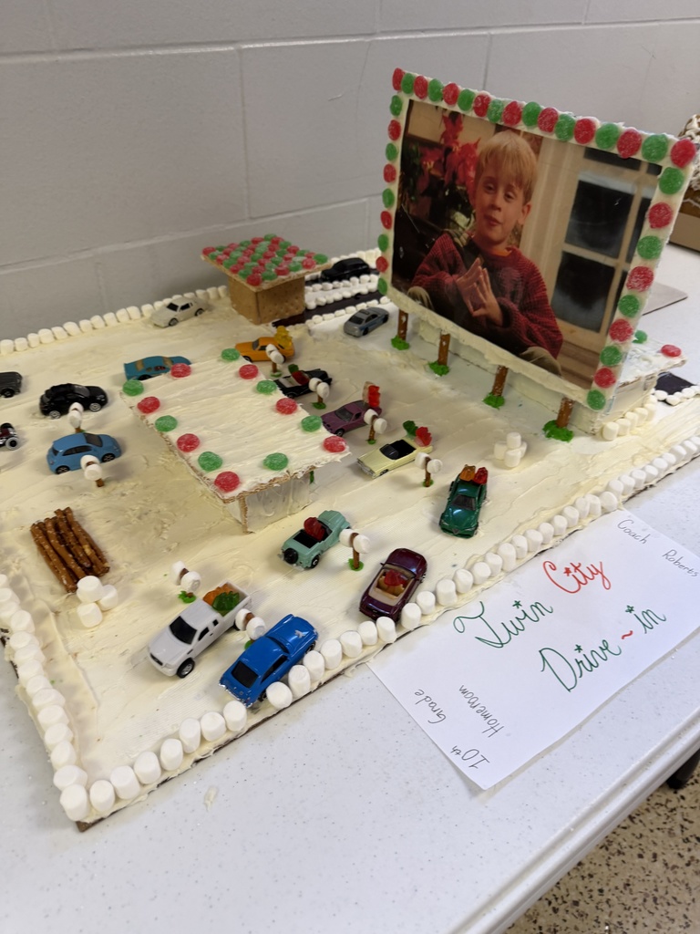 A collection of student-created gingerbread displays set on tables, featuring creatively decorated houses and scenes made from graham crackers, icing, candy, and small props. Designs include colorful candy roofs, snowy icing paths, miniature trees, a drive-in movie scene, a dinosaur-themed display, and humorous handwritten labels. The exhibits showcase imagination, craftsmanship, and festive holiday spirit.