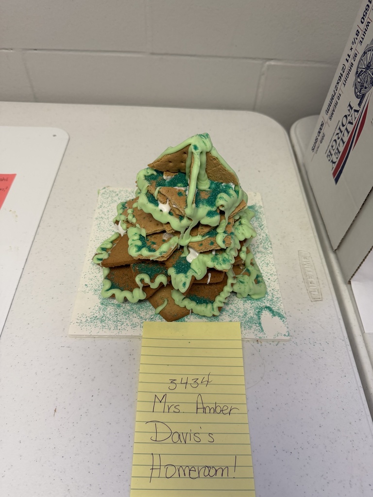 A collection of student-created gingerbread displays set on tables, featuring creatively decorated houses and scenes made from graham crackers, icing, candy, and small props. Designs include colorful candy roofs, snowy icing paths, miniature trees, a drive-in movie scene, a dinosaur-themed display, and humorous handwritten labels. The exhibits showcase imagination, craftsmanship, and festive holiday spirit.