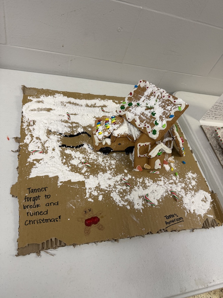 A collection of student-created gingerbread displays set on tables, featuring creatively decorated houses and scenes made from graham crackers, icing, candy, and small props. Designs include colorful candy roofs, snowy icing paths, miniature trees, a drive-in movie scene, a dinosaur-themed display, and humorous handwritten labels. The exhibits showcase imagination, craftsmanship, and festive holiday spirit.