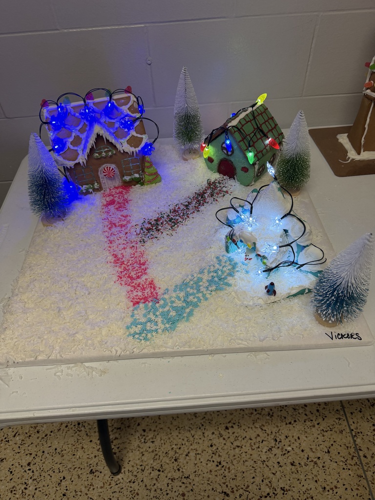 A collection of student-created gingerbread displays set on tables, featuring creatively decorated houses and scenes made from graham crackers, icing, candy, and small props. Designs include colorful candy roofs, snowy icing paths, miniature trees, a drive-in movie scene, a dinosaur-themed display, and humorous handwritten labels. The exhibits showcase imagination, craftsmanship, and festive holiday spirit.