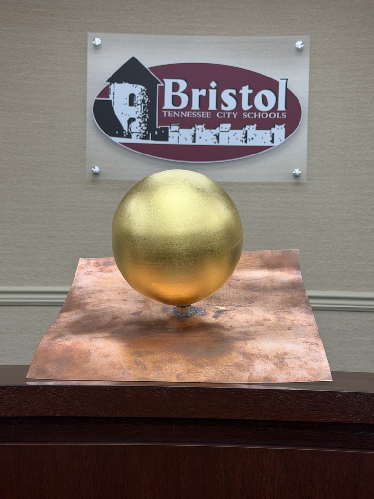 Gold-leafed copper finial ball mounted on a square copper base, displayed indoors on a wooden surface, with a Bristol Tennessee City Schools logo sign visible on the wall in the background.