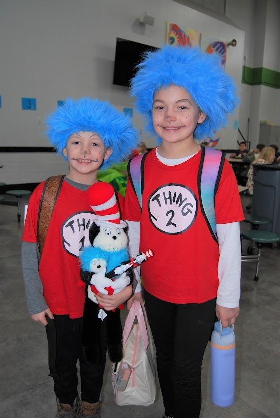 Two elementary students stand side by side in a school cafeteria dressed as Thing 1 and Thing 2 from Dr. Seuss. They wear bright red shirts labeled “Thing 1” and “Thing 2,” blue wigs, and playful face paint. One student holds a stuffed Cat in the Hat toy while they smile at the camera, with cafeteria tables and other students in the background.