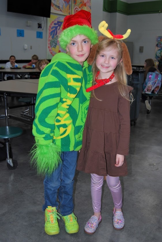 Two elementary students stand side by side in a school cafeteria wearing festive costumes. One is dressed as the Grinch in a green hoodie with furry cuffs and a Santa hat, and the other is dressed as Max the dog, wearing a brown outfit with floppy ears and a red collar. They smile at the camera with cafeteria tables and other students in the background.