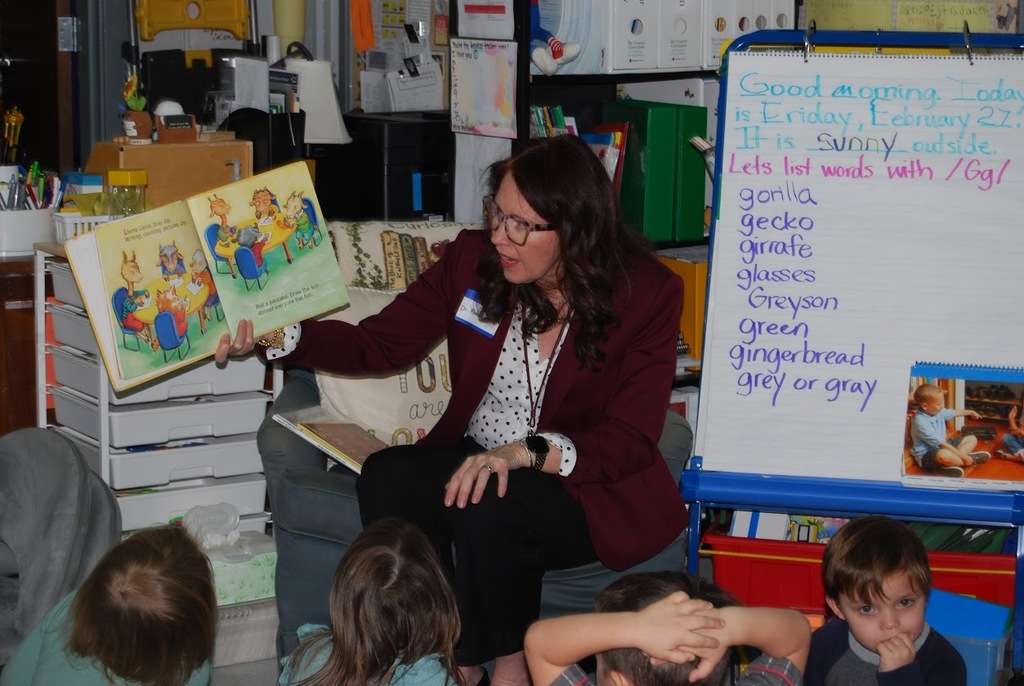 A guest reader sits in a classroom chair holding up a large picture book and reading to a small group of young students seated on the floor. A whiteboard beside her displays the date and a list of “G” words, including gorilla, gecko, giraffe, glasses, and gingerbread. Classroom shelves and supplies fill the background as the students look toward the book.