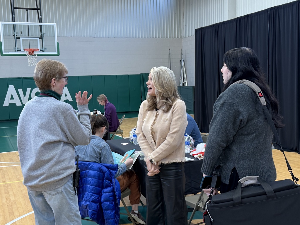 Three women stand talking and smiling in a school gymnasium during a community event. One gestures as she speaks while the others listen. Tables with water bottles, a basketball hoop, and green wall padding with “Avoca” lettering are visible in the background.