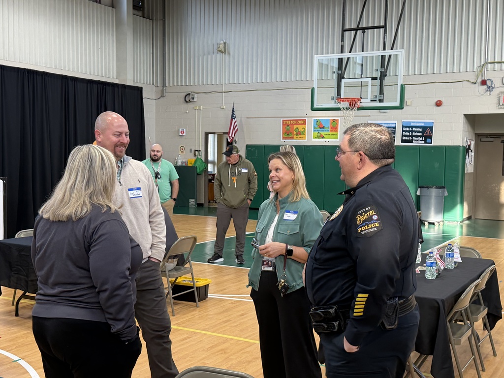 Four adults stand in a school gymnasium talking and smiling during a community event. A police officer in a Bristol, Tennessee uniform speaks with three other attendees wearing name tags. Tables with black tablecloths, a basketball hoop, and posters on the gym wall are visible in the background.