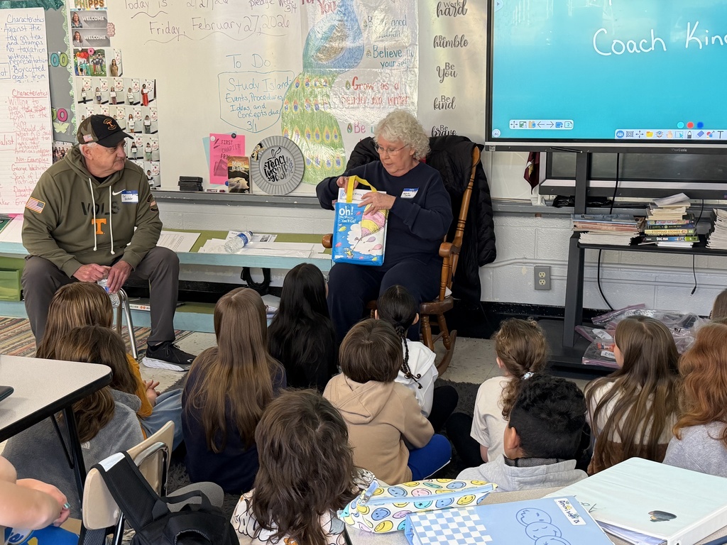 Two community guest readers sit at the front of a classroom speaking to a group of elementary students seated on the floor. One reader sits in a rocking chair holding a colorful book bag while addressing the students, and the other sits nearby listening. A whiteboard behind them shows the date and classroom notes, and a large digital screen is visible to the side.