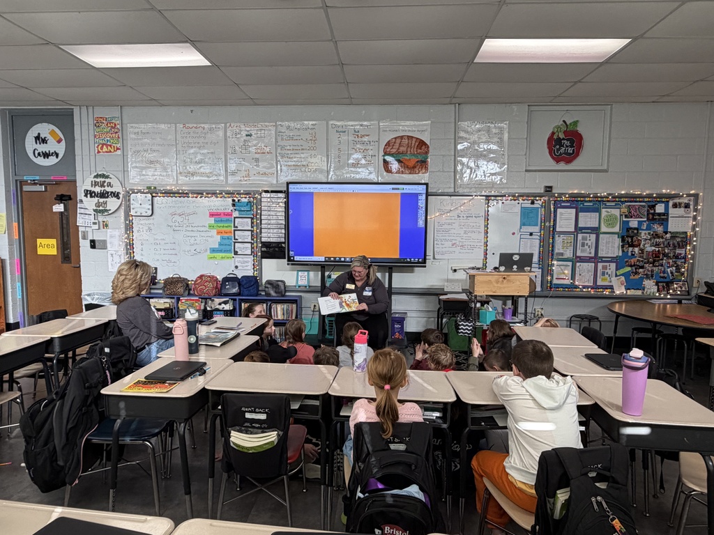 A guest reader sits at the front of an elementary classroom reading a picture book to a group of students gathered on the floor. Desks and backpacks fill the foreground, and classroom walls are covered with math anchor charts, posters, and a large digital display screen behind the reader.