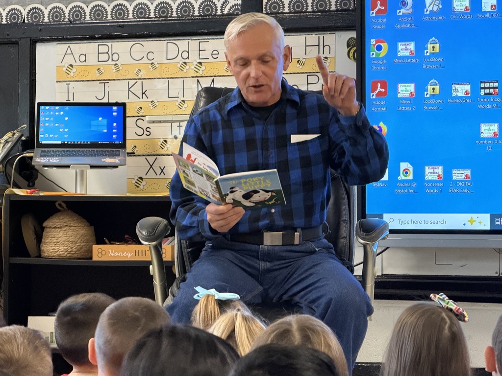 A community guest reader sits at the front of an elementary classroom reading from a children’s book while raising one finger for emphasis. Students sit on the floor facing him. An alphabet chart runs along the wall behind him, and a laptop and large digital screen are visible in the classroom.