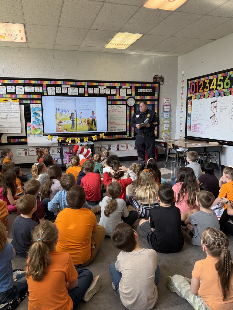 A Bristol, Tennessee police officer stands at the front of an elementary classroom reading to a large group of students seated on the floor. A colorful sound wall and number chart decorate the walls, and a digital display behind him shows an illustrated page from a children’s book. Students sit cross-legged and listen attentively.