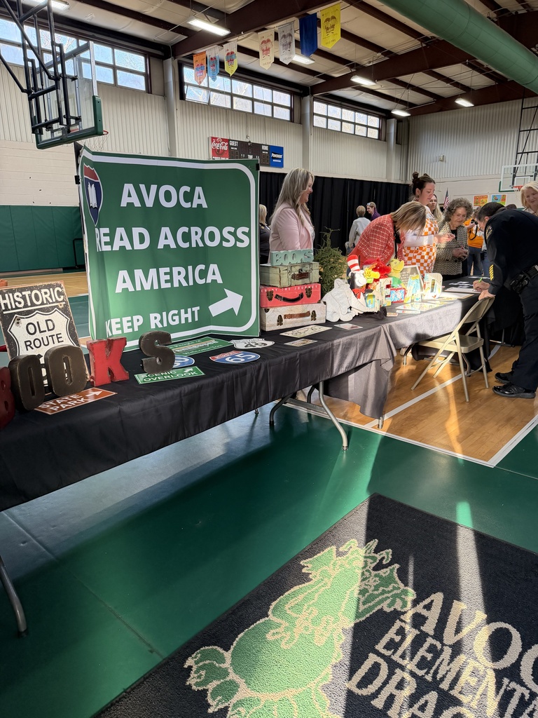 A guest reader sits in a classroom chair holding up a colorful picture book and reading to a small group of young students seated on the floor. A whiteboard beside her displays the date and a list of “G” words, including gorilla, gecko, giraffe, glasses, and gingerbread. The students look toward the reader as she shares the story.