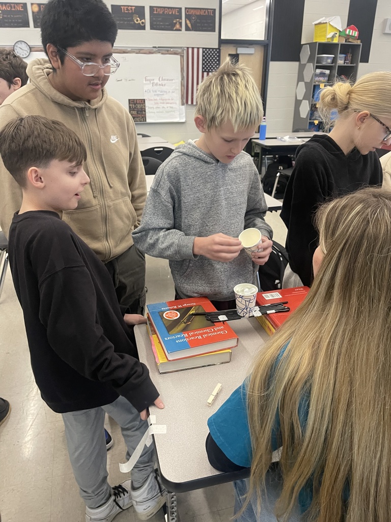 A group of Tennessee Middle School students gather around a table testing a small bridge structure made from black rods and tape, adding weight with stacked cups and books during an Engineers Week engineering challenge.