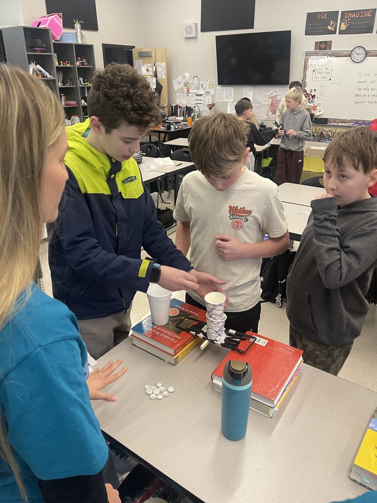 Students watch closely as one student carefully places stacked cups on a small bridge structure to test how much weight it can hold during a classroom engineering activity.