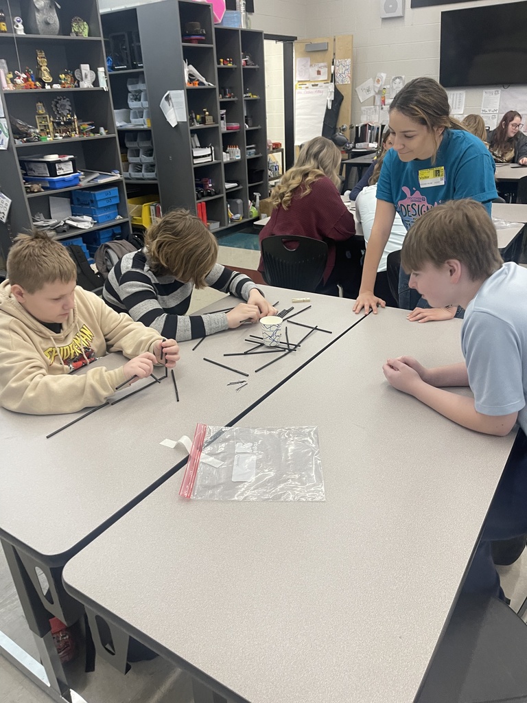 Three Tennessee Middle School students work together at a classroom table building a small bridge structure with black plastic rods while an engineer from Eastman Chemical Company observes and offers guidance during an Engineers Week design challenge.