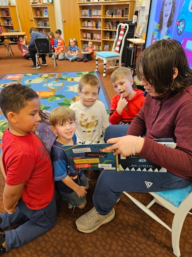 A Tennessee Middle School student reads a picture book to a small group of Fairmount Elementary students gathered around in the school library. The younger students stand and sit close by, looking at the open book as the middle school student points to the page during a Read Across America Week visit.