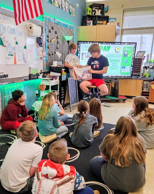 Two Tennessee Middle School students sit at the front of a Fairmount Elementary classroom reading a picture book to a group of younger students seated on a rug. One student holds the open book while reading aloud as the other stands nearby assisting, and the elementary students watch and listen during a Read Across America Week classroom visit.