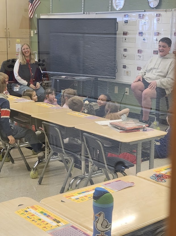 A Tennessee Middle School student sits at the front of a Fairmount Elementary classroom reading and talking with younger students gathered around their desks. A teacher sits nearby smiling as the elementary students listen and laugh during the Read Across America Week visit.