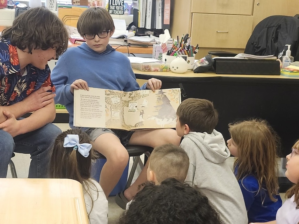 A Tennessee Middle School student holds open a picture book and reads aloud to a small group of Fairmount Elementary students seated on the floor in a classroom. Another middle school student sits nearby listening as the younger students look at the book during the Read Across America Week visit.