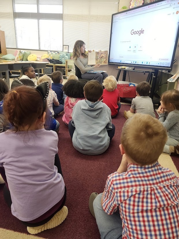 A Tennessee Middle School student reads a picture book aloud to a group of younger students seated on the floor during a classroom visit at Fairmount Elementary School for Read Across America Week. The elementary students sit closely together on a carpet listening to the story while the reader holds the book near a large interactive display.