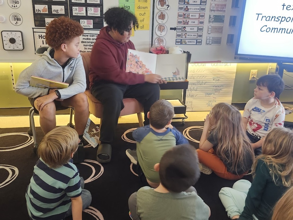Two Tennessee Middle School students sit at the front of a Fairmount Elementary classroom reading a picture book to a small group of young students seated on a patterned rug. One student holds the open book and reads while the other sits beside him holding another book, as the elementary students listen attentively during a Read Across America Week visit.