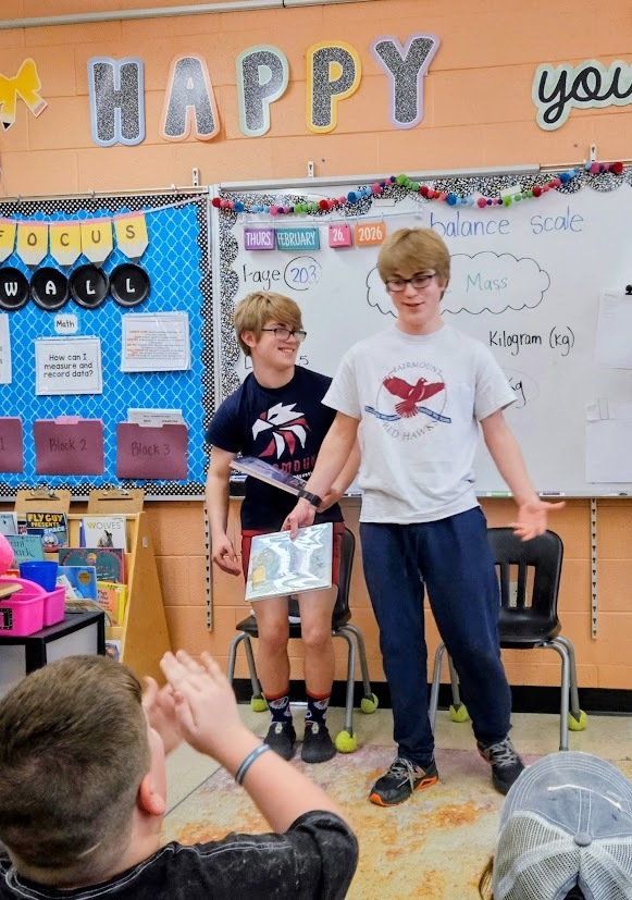 Two Tennessee Middle School students stand at the front of a Fairmount Elementary classroom after reading to younger students during Read Across America Week. One student holds a picture book while they speak to the class, and elementary students seated on the floor respond and ask questions.