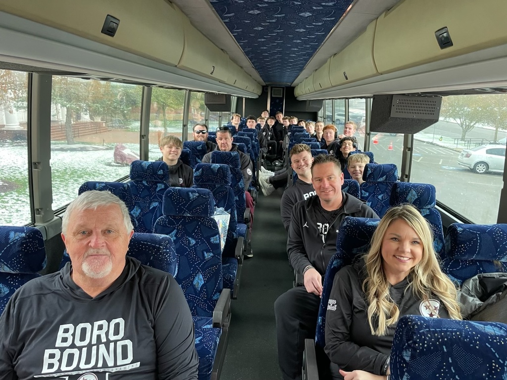 Inside a charter bus, Tennessee High School boys basketball players and coaches sit in blue patterned seats, smiling and looking toward the camera. A few coaches sit near the front, with players filling the rows behind them. Through the bus windows, snow can be seen on the ground outside as the team begins their trip to the state tournament.
