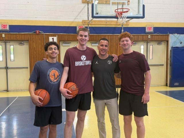 Four people stand together in a school gym, smiling at the camera. Three high school basketball players in athletic clothing hold basketballs while standing beside an adult staff member. A basketball hoop and gym doors are visible in the background.