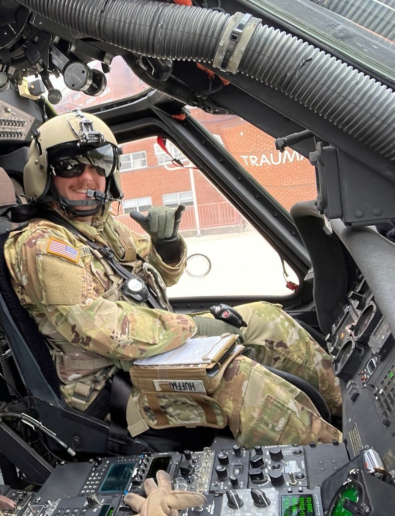 Interior view of a helicopter cockpit with control panels and instruments visible, along with part of a pilot’s gloved hand. The top of the image shows the helicopter’s frame and components, with a partial view of a building outside. The image appears to be a screenshot from a messaging app, with portions of text messages overlaying the scene.