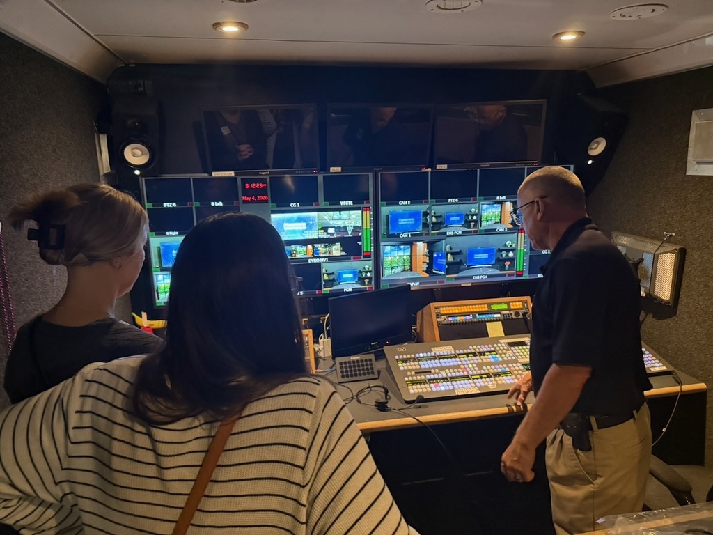 Three adults stand in a television control room, looking at multiple monitors displaying live video feeds. A staff member gestures toward a large production switcher covered in colorful buttons, explaining how the equipment works while the others observe.