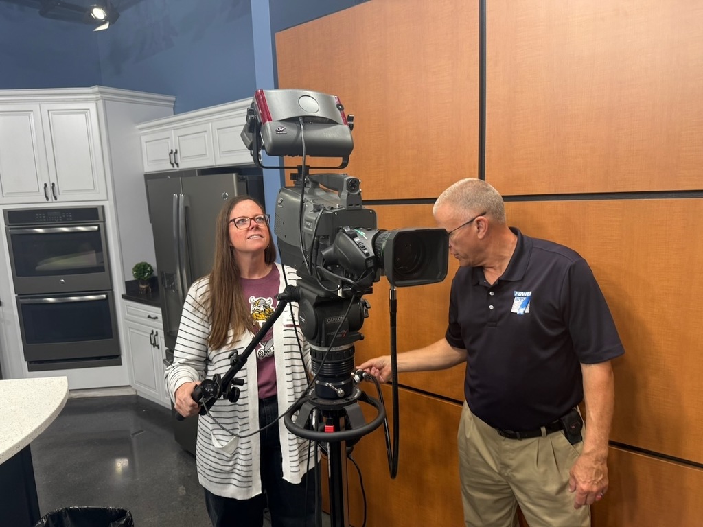 Two adults stand beside a professional video camera mounted on a tripod in a studio kitchen setting. One person adjusts the camera controls while the other looks up at the camera, smiling slightly. Behind them are white cabinets, a stainless steel refrigerator and ovens, and wood-paneled walls, suggesting a media or production space.